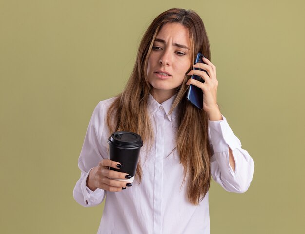 Chica bastante caucásica joven despistada sostiene la taza de papel y habla por teléfono