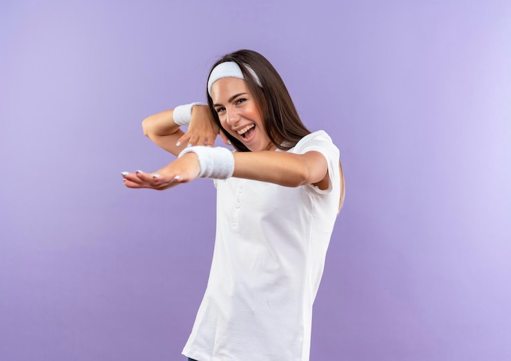 Foto gratuito chica bastante deportiva alegre con diadema y muñequera estirando la mano con otra cerca de la cabeza aislada en la pared púrpura