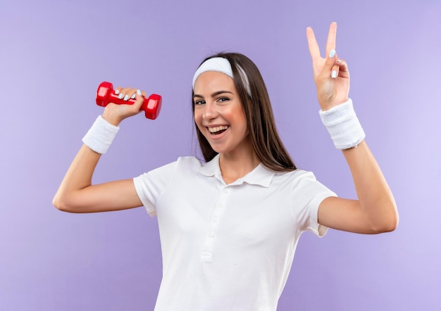 Chica bastante deportiva alegre con diadema y muñequera sosteniendo pesas haciendo el signo de la paz aislado en la pared púrpura