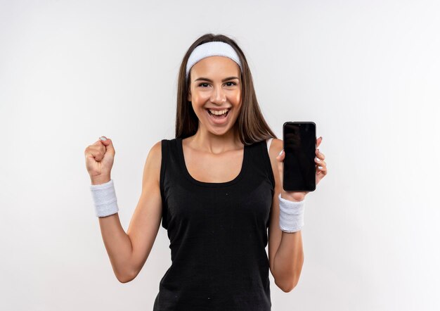 Chica bastante deportiva alegre con diadema y muñequera sosteniendo el teléfono móvil y levantando el puño aislado en la pared blanca