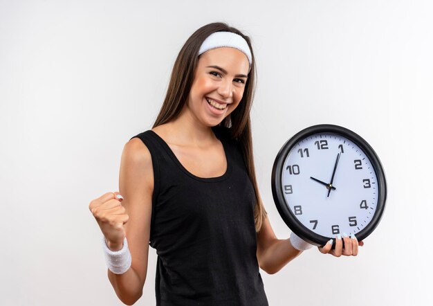 Chica bastante deportiva alegre con diadema y pulsera sosteniendo el reloj con el puño cerrado aislado en la pared blanca