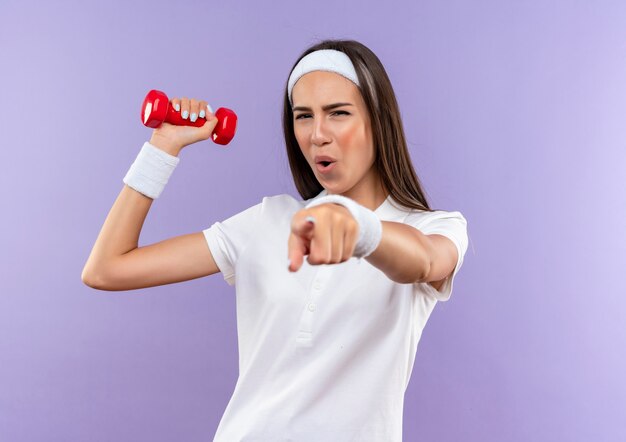 Chica bastante deportiva confiada con diadema y muñequera sosteniendo mancuernas apuntando aislado en la pared púrpura