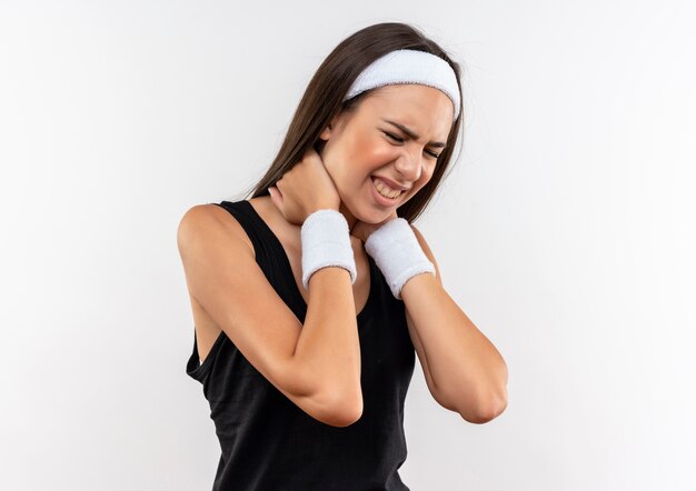 Chica bastante deportiva dolorida con diadema y muñequera poniendo las manos en el cuello con los ojos cerrados aislado en la pared blanca