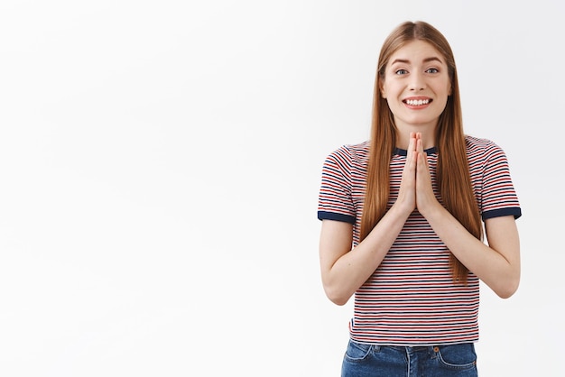 Chica bastante guapa con camiseta a rayas agradeciendo a un amigo por ayudar a tomarse de la mano en oración rogando favor sonriendo alegremente juntando las manos como pegajoso pidiéndole a papá que le preste un auto de fondo blanco