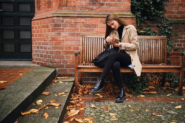 Chica bastante informal con mochila buscando tarjeta en billetera sentada en un banco en la calle de la ciudad