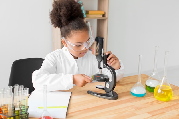 Chica en bata de laboratorio y gafas de seguridad mirando a través del microscopio