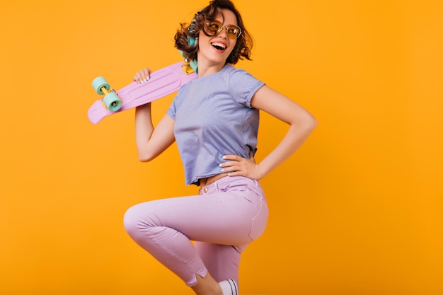 Chica bien formada activa con longboard rosa posando con una sonrisa alegre Foto interior de una mujer caucásica relajada con gafas de color amarillo bailando en el estudio