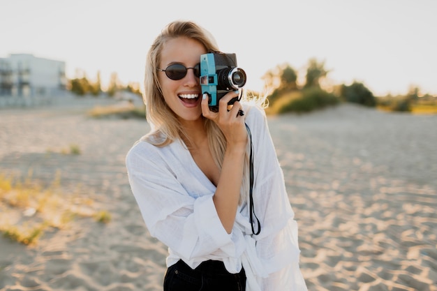 Chica bien formada con estilo con cámara retro posando en la playa soleada. Vacaciones de verano. Estado de ánimo tropical. Concepto de libertad y viaje.