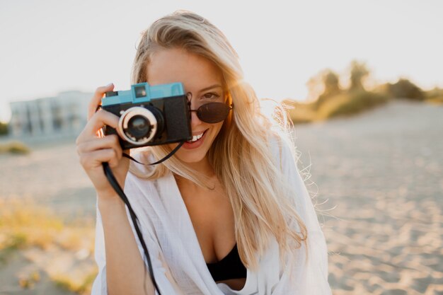 Chica bien formada con estilo con cámara retro posando en la playa soleada. Vacaciones de verano. Estado de ánimo tropical. Concepto de libertad y viaje.