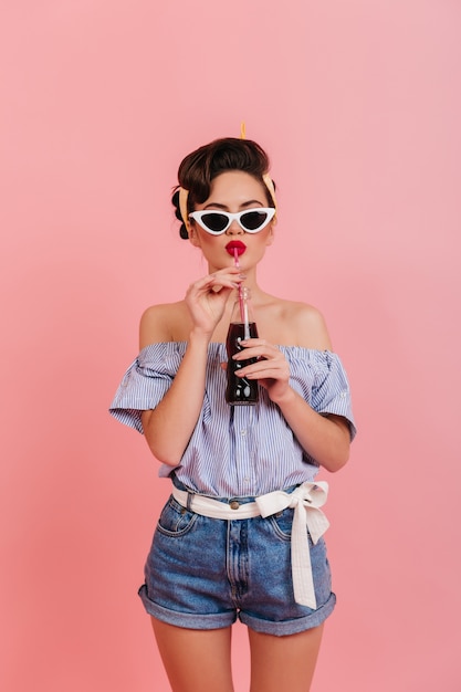 Foto gratuita chica bien formada en pantalones cortos de mezclilla y blusa a rayas bebiendo bebidas. foto de estudio de dama pinup en gafas de sol.