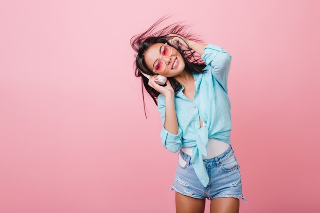 Chica bien formada en pantalones cortos de mezclilla de moda escuchando música con una sonrisa y tocando grandes auriculares. Retrato de interior de hermosa mujer hispana en camisa de algodón feliz bailando.