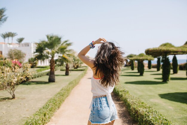 Chica bien formada de pelo largo con camisa blanca caminando por el callejón de las palmeras bajo un cielo azul en la mañana soleada. Retrato de elegante joven divertida bailando en el parque en resort en vacaciones de verano.