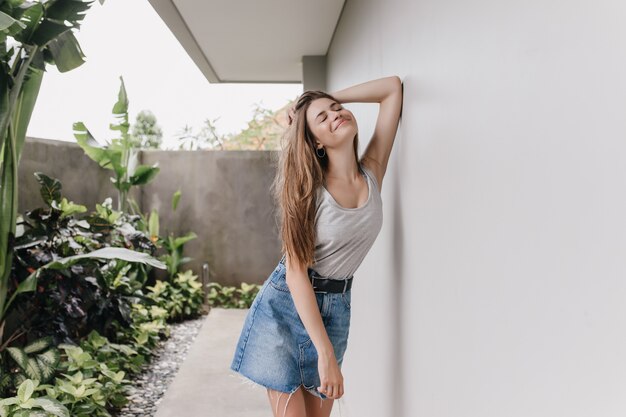 Chica bien formada de pelo oscuro en traje casual de verano posando al aire libre junto a las plantas. Retrato de mujer encantadora en falda de mezclilla de pie junto a la pared con los ojos cerrados.