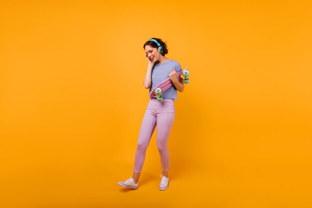 Chica bien formada en ropa casual colorida escuchando música en auriculares. Modelo de mujer alegre con corte de pelo corto con patineta.