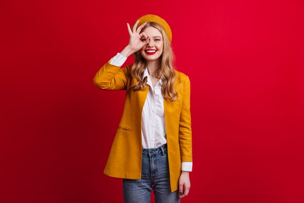 Chica bien vestida con boina mostrando signo bien. modelo femenino en chaqueta amarilla aislada en la pared roja.