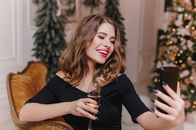 Chica bien vestida sosteniendo un teléfono negro y tomando una foto de sí misma. Encantadora mujer de cabello oscuro con copa de vino con smartphone para selfie con árbol de Navidad en la pared.