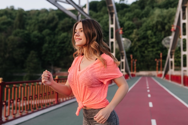 Chica blanca despreocupada en camiseta rosa corriendo. Foto exterior de la elegante dama europea entrenando en la mañana.