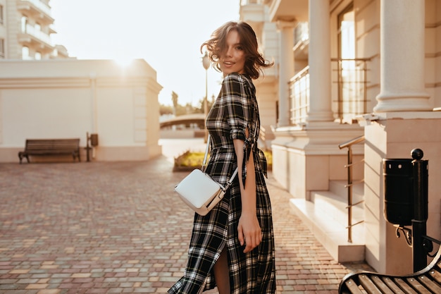 Chica blanca elegante con bolso pequeño mirando hacia atrás en la calle. Modelo de mujer bastante rizado en vestido largo caminando por la ciudad en día de otoño.