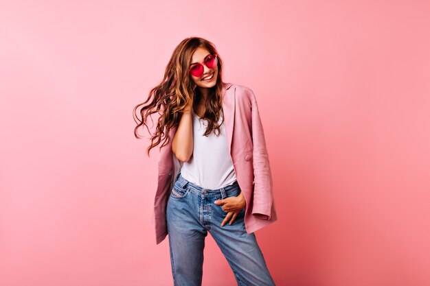 Chica blanca emocionada en gafas con estilo brillantes posando en rosa. Mujer rizada soñadora jugando con su pelo pelirrojo y riendo.