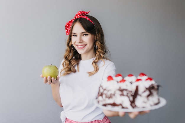 Chica blanca de ensueño sosteniendo gran pastel de cumpleaños con bayas y sonriendo. La atractiva modelo femenina de cabello oscuro no puede decidir qué elegir entre tarta y manzana.