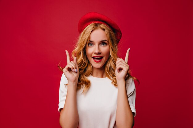 Chica blanca extasiada en boina posando con asombro. Elegante modelo de mujer caucásica en camiseta de pie en la pared roja.