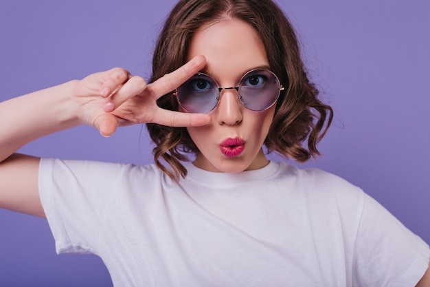 Chica blanca guapa lleva camiseta posando con el signo de la paz en estudio. Modelo de mujer morena alegre en gafas de sol divirtiéndose en la pared púrpura.