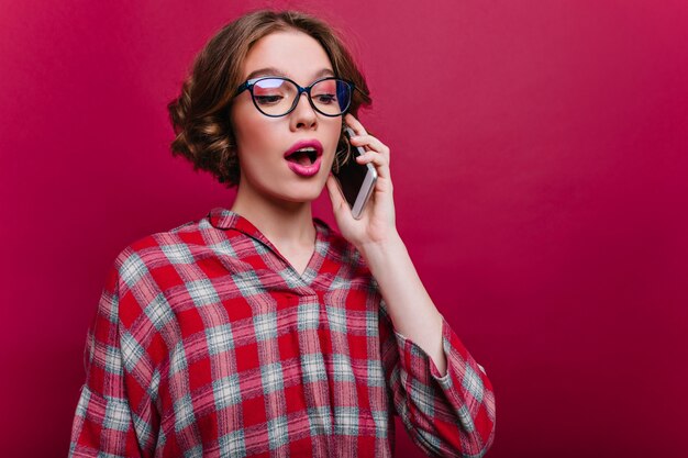 Chica blanca interesada con maquillaje rosa brillante hablando por teléfono en la pared clarete. Mujer joven guapa con smartphone.