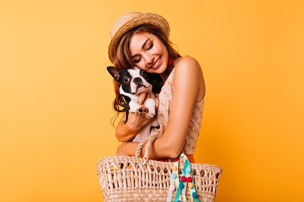 Foto gratuita chica blanca pelirroja posando en estudio con su mascota. atractiva señorita con sombrero con bulldog francés en brillante.