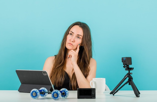 Chica blogger está pensando sosteniendo el dedo en la mejilla frente a la cámara sobre fondo azul.