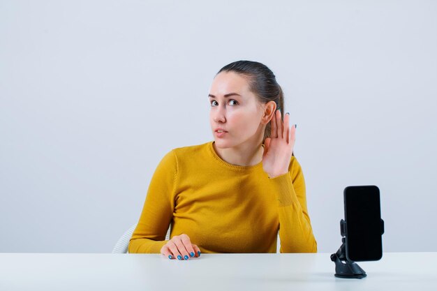 Chica blogger está sosteniendo la mano detrás de la oreja sentada frente a la cámara móvil sobre fondo blanco.