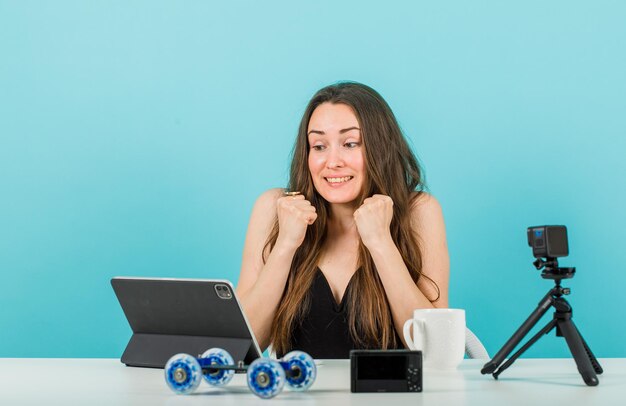Chica blogger está sosteniendo puños en el pecho mirando la tableta sobre fondo azul.