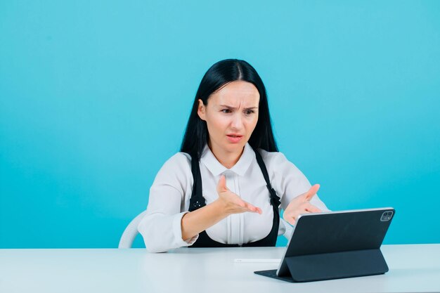 Chica bloguera enojada está extendiendo las manos a la pantalla de la tableta con fondo azul