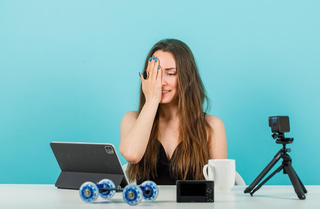 Chica bloguera está mirando la tableta cubriendo la mitad de la cara con la mano sobre fondo azul