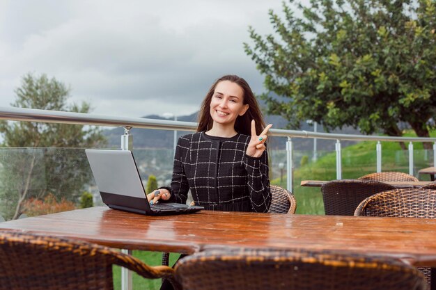 Chica bloguera con laptop muestra un gesto de victoria a la cámara sentándose en el fondo de la vista de la naturaleza