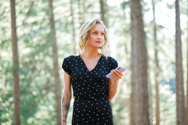 Chica bloguera rubia está mirando awya sosteniendo el móvil en el fondo de la naturaleza