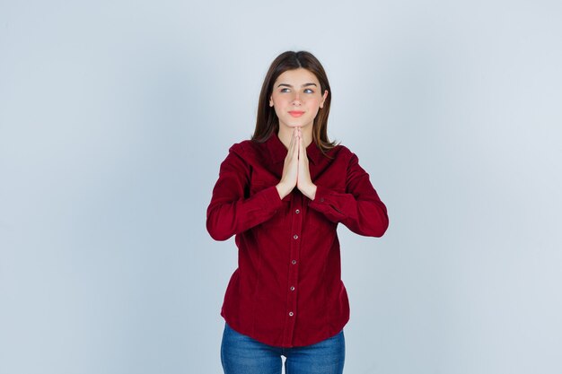 Chica en blusa burdeos manteniendo las manos en gesto de oración, mirando a un lado y mirando agradecido