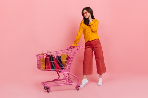 Chica en blusa naranja y gafas de sol con sonrisa mira muchas de sus compras en carro rosa del supermercado.