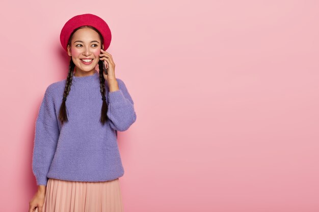 Chica bonita habladora con apariencia oriental disfruta de una conversación telefónica, tiene un teléfono celular moderno cerca de la oreja, usa una elegante boina roja y un suéter violeta