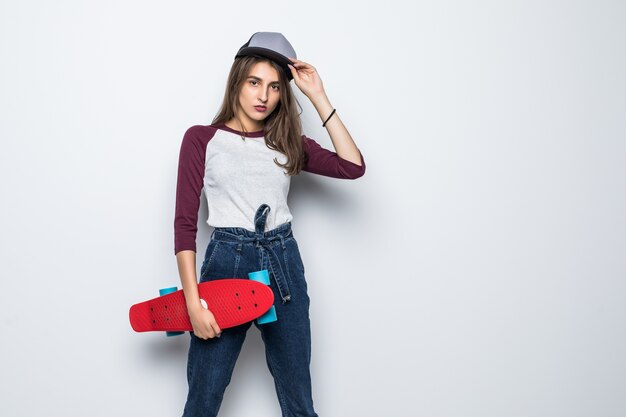 Chica bonita patinadora sosteniendo patineta roja en sus manos aisladas en la pared blanca