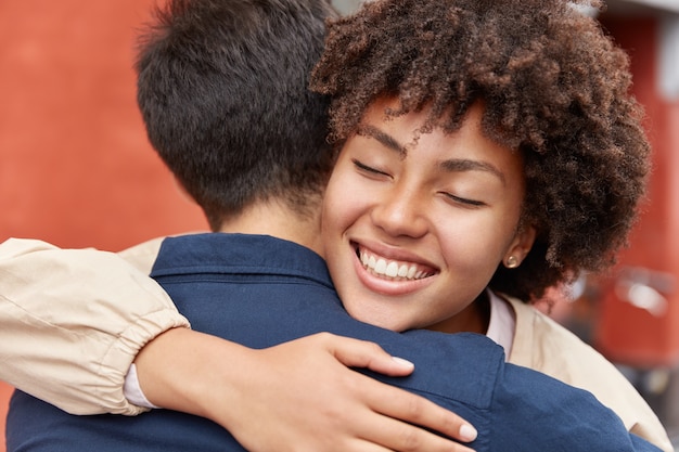 La chica bonita de piel oscura complacida tiene una sonrisa con dientes, mantiene los ojos cerrados, abraza a su viejo amigo, tiene una buena relación