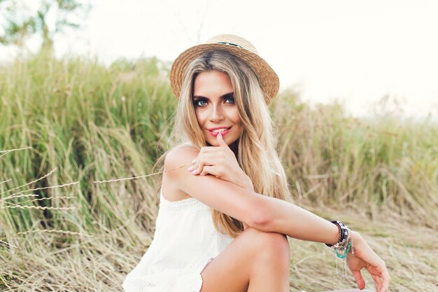 Chica bonita rubia con pelo largo está posando para la cámara en el fondo del campo. Lleva sombrero, vestido blanco y sonríe a la cámara.