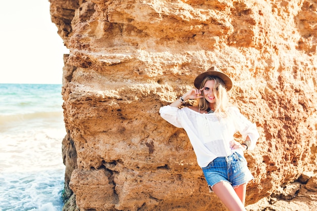 Chica bonita rubia con el pelo largo está posando para la cámara en la playa sobre fondo de piedra. Ella esta sonriendo.