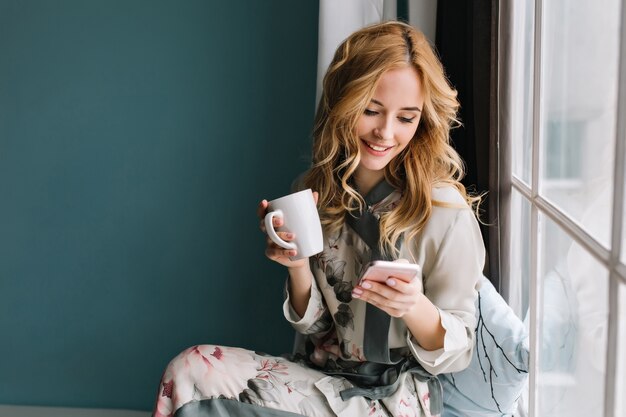 Chica bonita rubia sentada en el alféizar de la ventana con una taza de café, té y smartphone en las manos. Tiene el pelo largo y rubio ondulado, sonríe y mira su teléfono. Usando un hermoso pijama de seda.