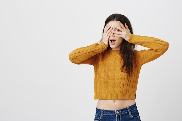 Foto gratuita chica bonita sorprendida y sorprendida se cubre los ojos con las manos, pero mira a través de los dedos con curiosidad