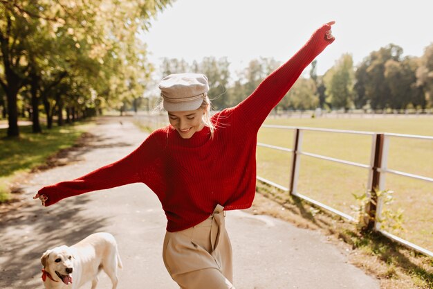 Chica brillante divirtiéndose con su perro en el parque de otoño. Sonriente mujer rubia bailando en tiempo soleado con su labrador.