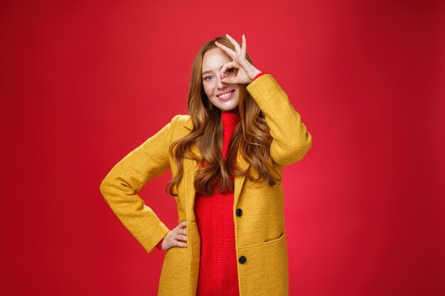 Chica brillante y soleada que se siente feliz incluso en los días de lluvia de pie con un elegante abrigo amarillo sobre fondo rojo que muestra un buen gesto sobre los ojos, sonriendo ampliamente a la cámara, sosteniendo la mano en la cintura.