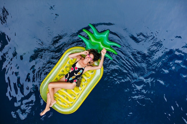 Chica bronceada en divertido traje de baño escalofriante en el balneario. Retrato de arriba de la risa modelo bastante femenino acostado en el colchón.