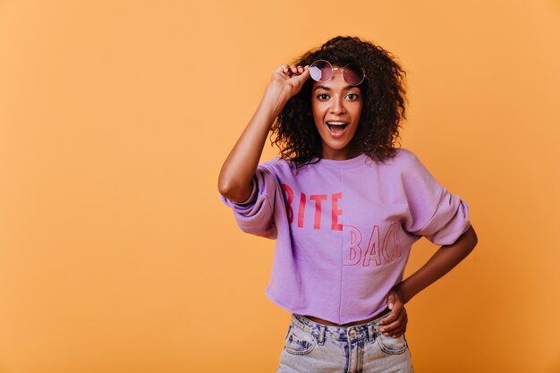 Chica de buen humor asombrada posando en naranja. Encantadora dama africana rizada