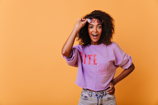 Chica de buen humor asombrada posando en naranja. Encantadora dama africana rizada