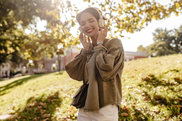 Foto gratuita chica de buen humor con chaqueta blanca con bolso escuchando música al aire libre mujer fresca con cabello corto en auriculares sonriendo afuera